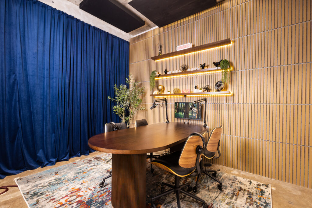 Wide view of a Band Barracks podcast studio 109 in Houston, featuring a wooden recording table, boom-mounted microphones, headphones, and seating. The room includes blue curtains, wood-slat acoustic walls, warm LED-lit shelves, a monitor, and decorative plants.