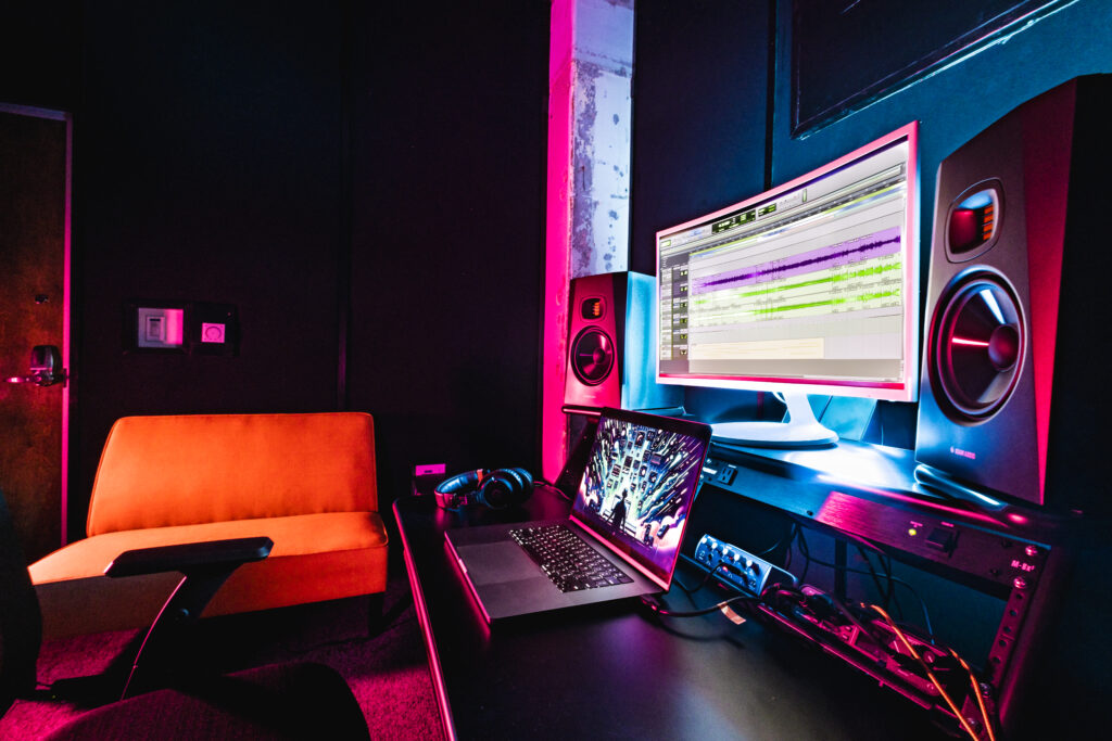 Band Barracks Recording Studio 107 with a computer running audio recording software, studio monitors, laptop, headphones, audio equipment, and lounge seating under blue and magenta lighting.
