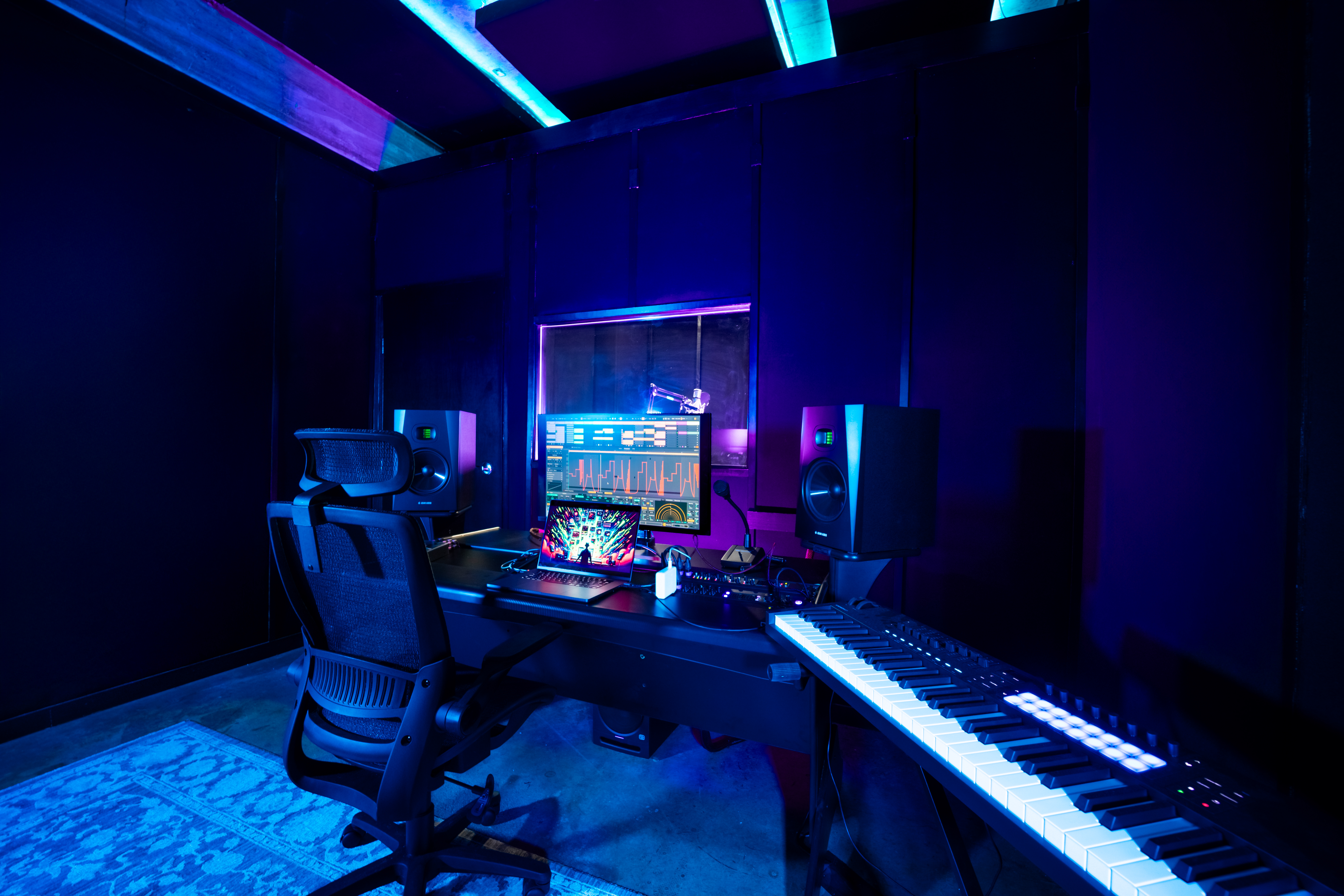 Band Barracks recording studio workspace with studio monitors, a computer and laptop running audio production software, a MIDI keyboard, and a microphone visible through the booth window under blue and purple lighting.