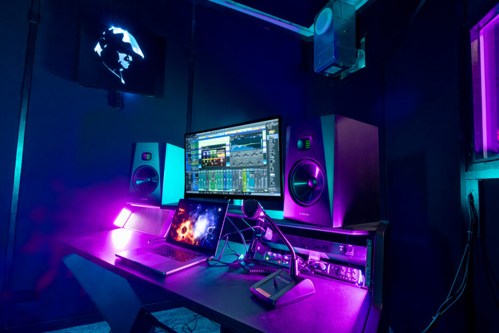Band Barracks recording studio workspace with a computer running audio production software, studio monitors, laptop, microphone, and recording equipment under blue and magenta lighting.