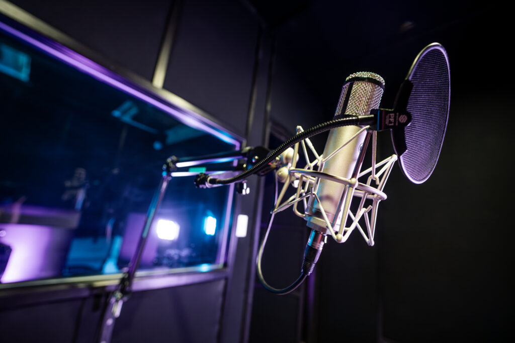 Close-up of a condenser microphone with a pop filter and shock mount inside a Band Barracks recording studio, with the studio window and blue-purple lighting in the background.
