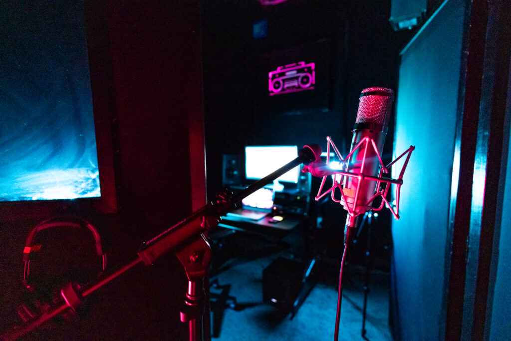 Condenser microphone in a shock mount inside a Band Barracks recording studio, with a computer workstation visible in the background under blue and magenta lighting.