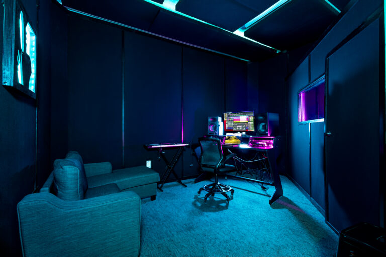 Wide view of a Band Barracks recording studio with a computer workstation, studio monitors, MIDI keyboard, office chair, couch, and acoustic treatment under blue and magenta lighting.
