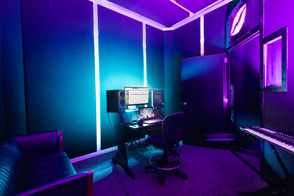 Band Barracks recording studio with a computer workstation, studio monitors, laptop, MIDI keyboard, office chair, and couch under blue and purple lighting.