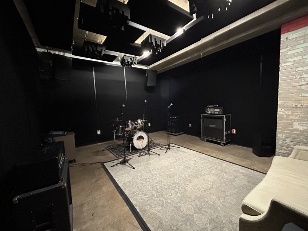 Band Barracks Rehearsal Studio 123, the facility’s largest rehearsal room, featuring a drum kit, multiple microphone stands, amplifiers, acoustic ceiling panels, a large area rug, and lounge seating.