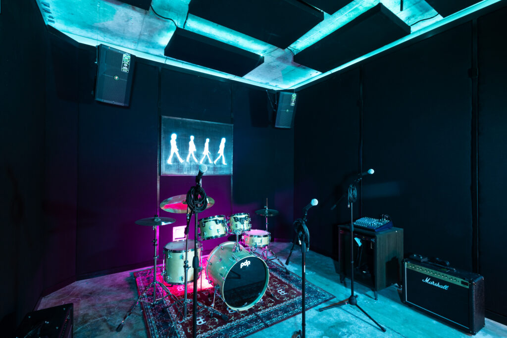 Band Barracks rehearsal studio with a PDP drum kit, multiple microphone stands, a Marshall amplifier, mixer, wall-mounted speakers, and acoustic ceiling panels under blue and magenta lighting.