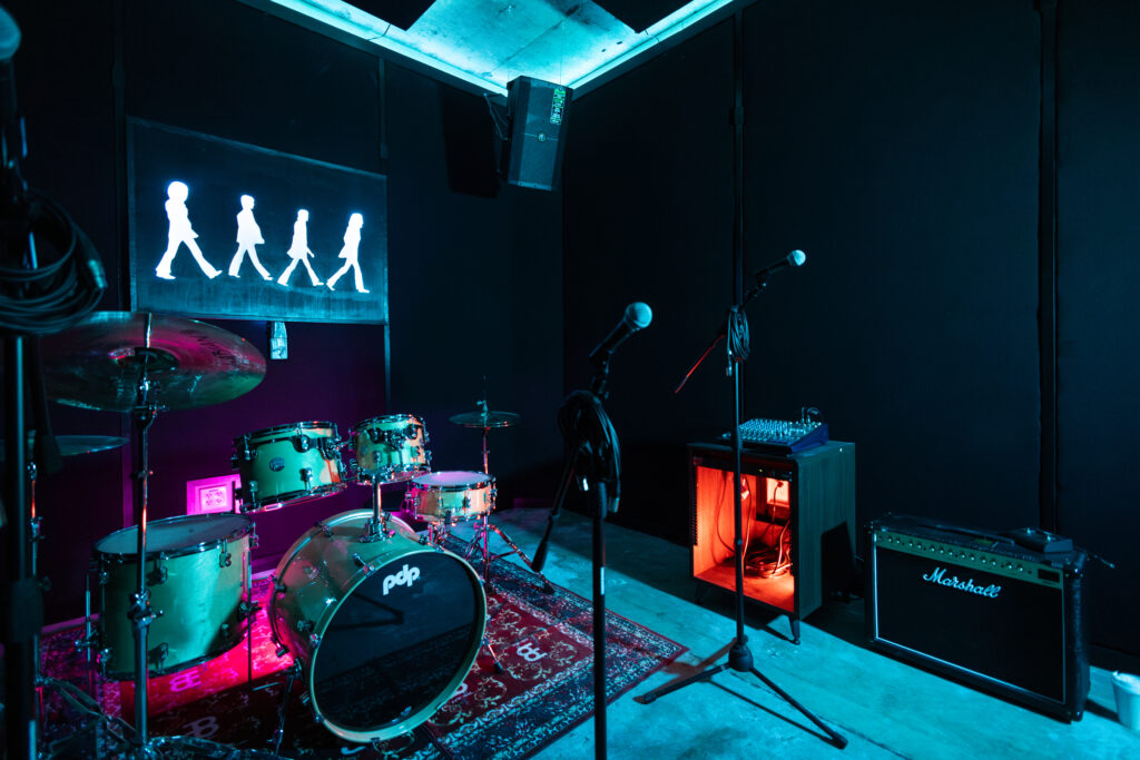 Band Barracks rehearsal studio with a PDP drum kit, multiple microphone stands, a Marshall amplifier, mixer, wall-mounted speaker, and acoustic treatment under blue and magenta lighting.
