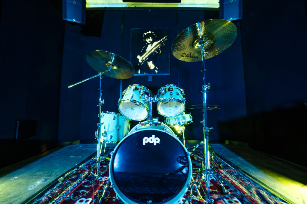 PDP drum kit with large cymbals set up on a patterned rug inside a Band Barracks rehearsal studio, with wall-mounted speakers and blue-green studio lighting.